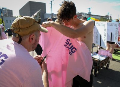 (L–R) Alexander Ortega scissor-slices a parade T-shirt for Jace Burbidge. SLUG ran out of smaller sizes, so XL shirts were "made" to fit. Photo: John Barkiple