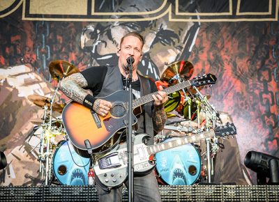 Wielding two guitars, Volbeat lead singer Michael Poulsen sings as he picks out an acoustic melody.