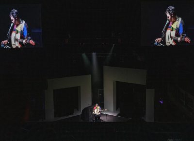 Marinelli playing on stage with giant white arches behind him and his performance also being casted on to the monitors above for the audience.