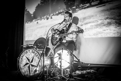 Demonstrating his talent as a mono band, Jacob T. Skeen sings in front of a projection of a cemetery, skillfully playing both drums and guitar.