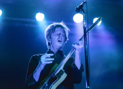 Britt Daniel playing guitar on stage with blue lighting.