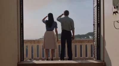Two people stand looking out a balcony, their backs to the camera.