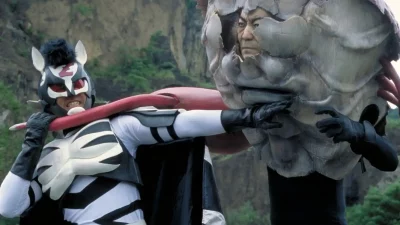 A man in a zebra-like costume attempts to fend off another man in costume. The scene is absurd.