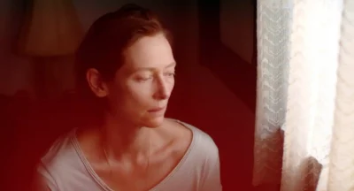 A woman (Tilda Swinton) looks out a window with translucent curtains. She looks sullen.