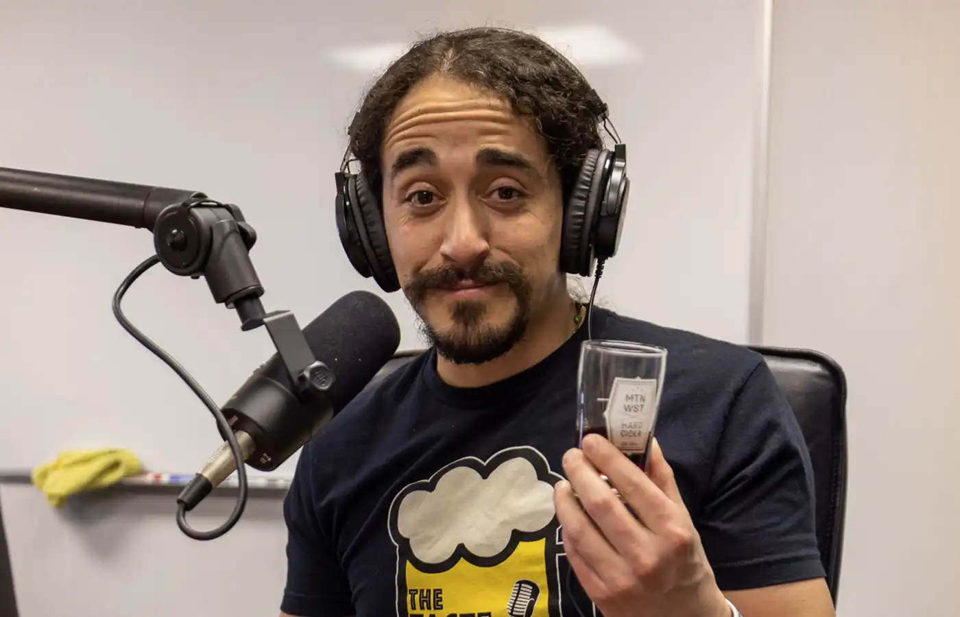 David Jimenez holds up a tasting glass and looks at the camera. A microphone encroaches out of frame, near his head. He has a pensive look with one eyebrow raised. Photo: John Barkiple