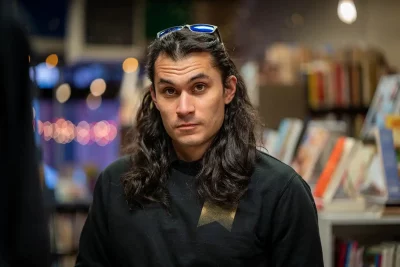 Danny Patiño looks at the camera seriously against a library backdrop. He is wearing a black sweater.