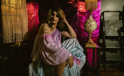 Tara Lipsyncki sits in a billowy pink-and-blue outfit in front of a pink set designed by Bonneville Jones. Two lamps bookend her on either side. 