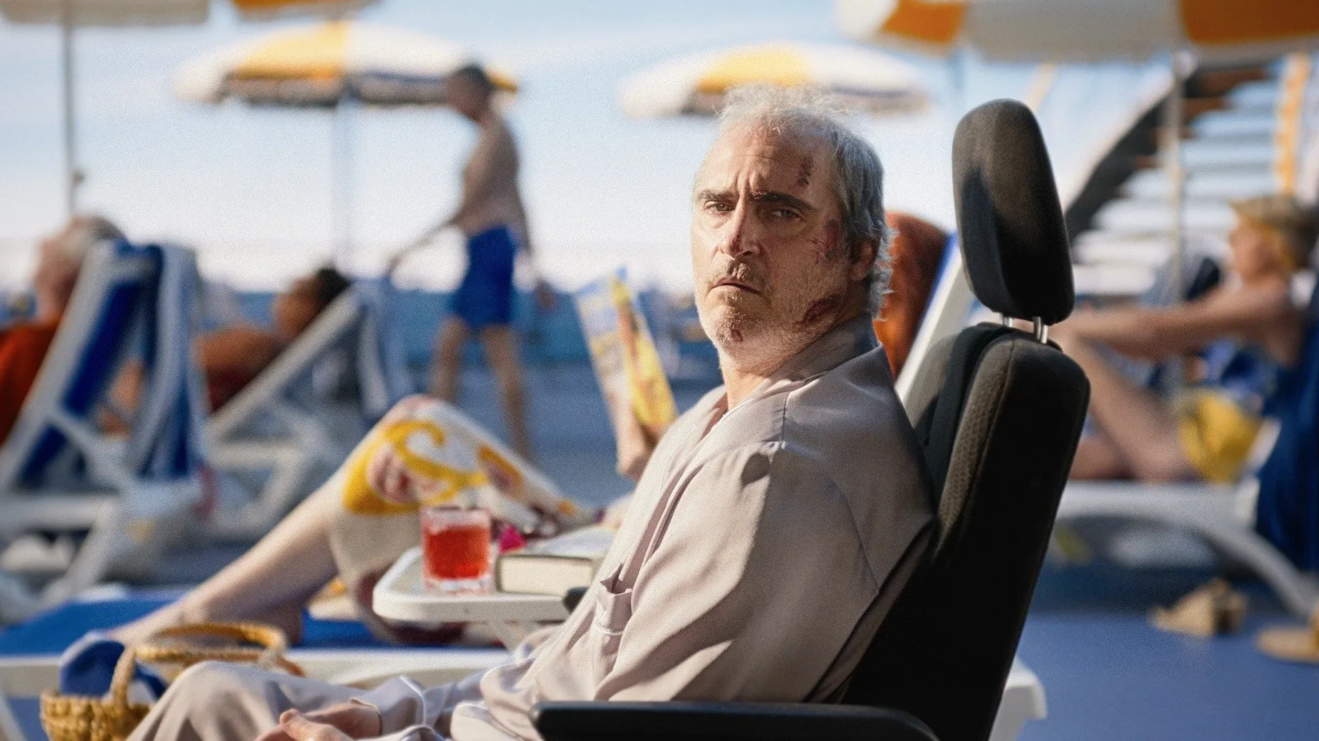 Joaquin Phoenix sits on a chair among people sitting on beach chairs.