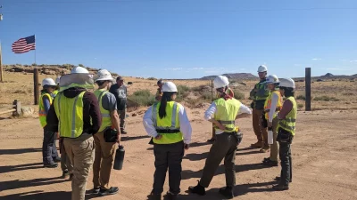 A group of DesignBuildBLUFF students in neon-yellow construction gear stand around a housing plot.