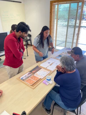 Around a large wooden table, four DesignBuildBLUFF students discuss building plans.