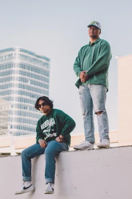 Bobby B Mac sits and Vehnu Moon stands on a ledge in front of a cityscape.