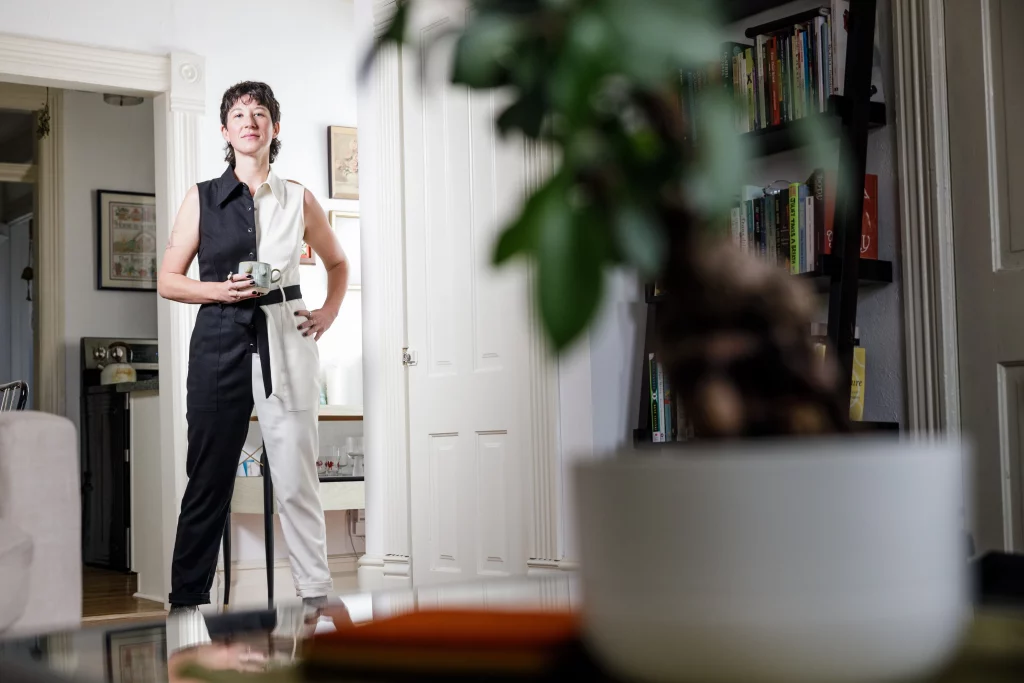 Amanda Madden stands in their home, one hand holding a coffee mug and the other on their hip. They are wearing a black and white sleeveless jumpsuit.