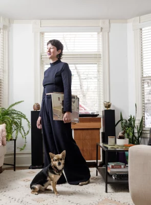 Amanda Madden stands in a living room in an all-black outfit, looking to the side, with their dog sitting in front of them.