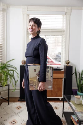 Amanda Madden stands in front of a window wearing an all-black outfit, holding records and smiling.