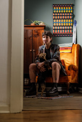 Amanda Madden sits on an orange velvet chair, wearing platform Doc Martens and holding their dog.