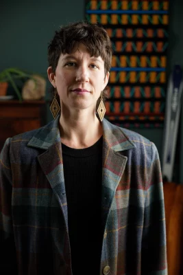 Amanda Madden stands in front of a colorful tapestry, wearing large gold earrings and a red and green plaid blazer.