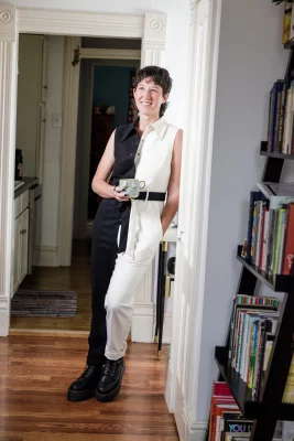 Amanda Madden leans against a wall, wearing a half-black-half-white jumpsuit and holding a coffee mug.