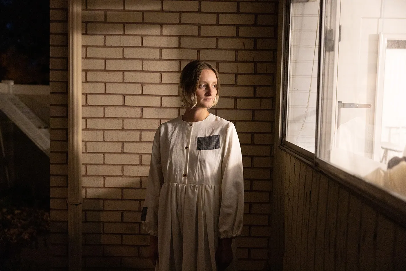 Local Designer Abigail Rue Jenson stands wearing a piece from her "Patched Up" collection against a brick background. Light from her home shines over her.