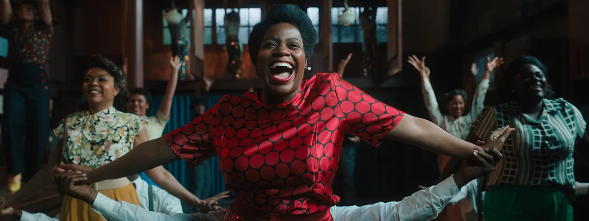 A woman in bright red dress surrounded by background dancers belts out lyrics in the middle of a musical number.