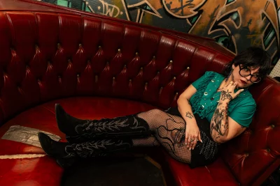 Kat Aleman leans on a red leather couch wearing black cowboy boots and a teal tassled top.