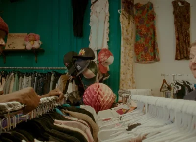 A young African-American man and an older Caucasian mad exchange glances over two stuffed clothing racks in a clothing store.
