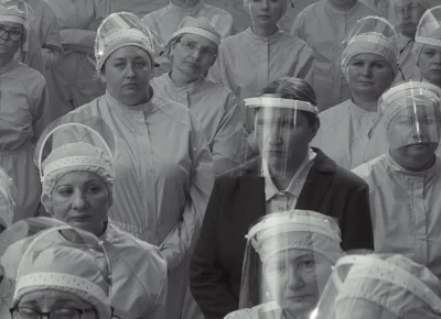 A black and white image of a woman in a black pantsuit in a sea of other women in greyscale scrubs faces the opposite way of them.