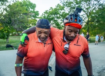 A younger son has his arm around his older father while both are dressed in bike racing gear.