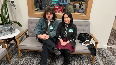 Parviz Faiz and Bianca Velasquez sit on a grey coach in front of a recording booth. 