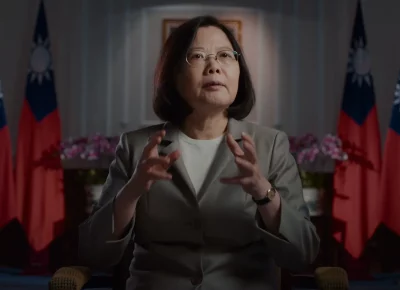 A bespectacled woman with short brown hair in a beige pantsuit is in mid-sentence during an interview.