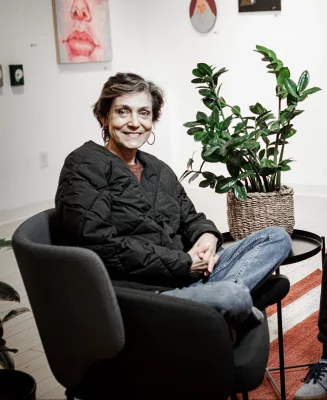 Betsy L Ross sits in a chair, wearing a coat and smiling at the camera.