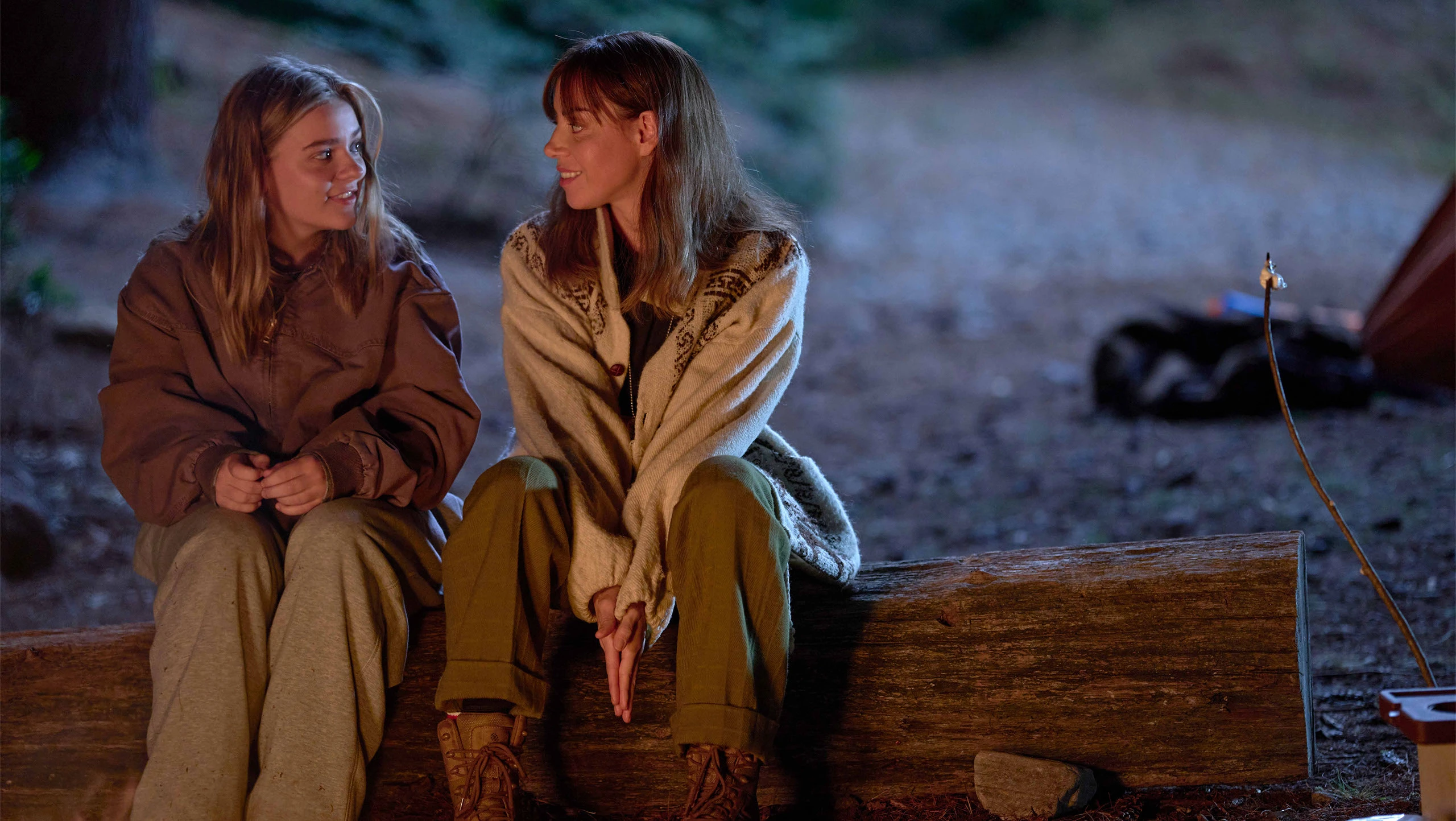 Two women sit on a log, looking at each other.