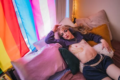 Robin D'Banc lays on a pile of pillows with the LGBTQ+ and transgender Pride flags behind her.