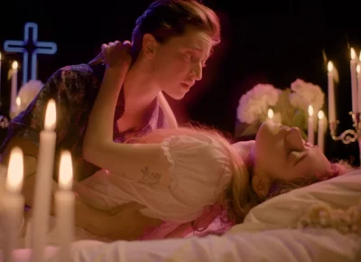 A young couple embraces on a white bed, in a cathedral setting, is surrounded by candles and neon crosses.