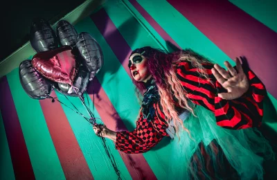 Shortcake the Clown holds balloons and leans against a striped wall. 