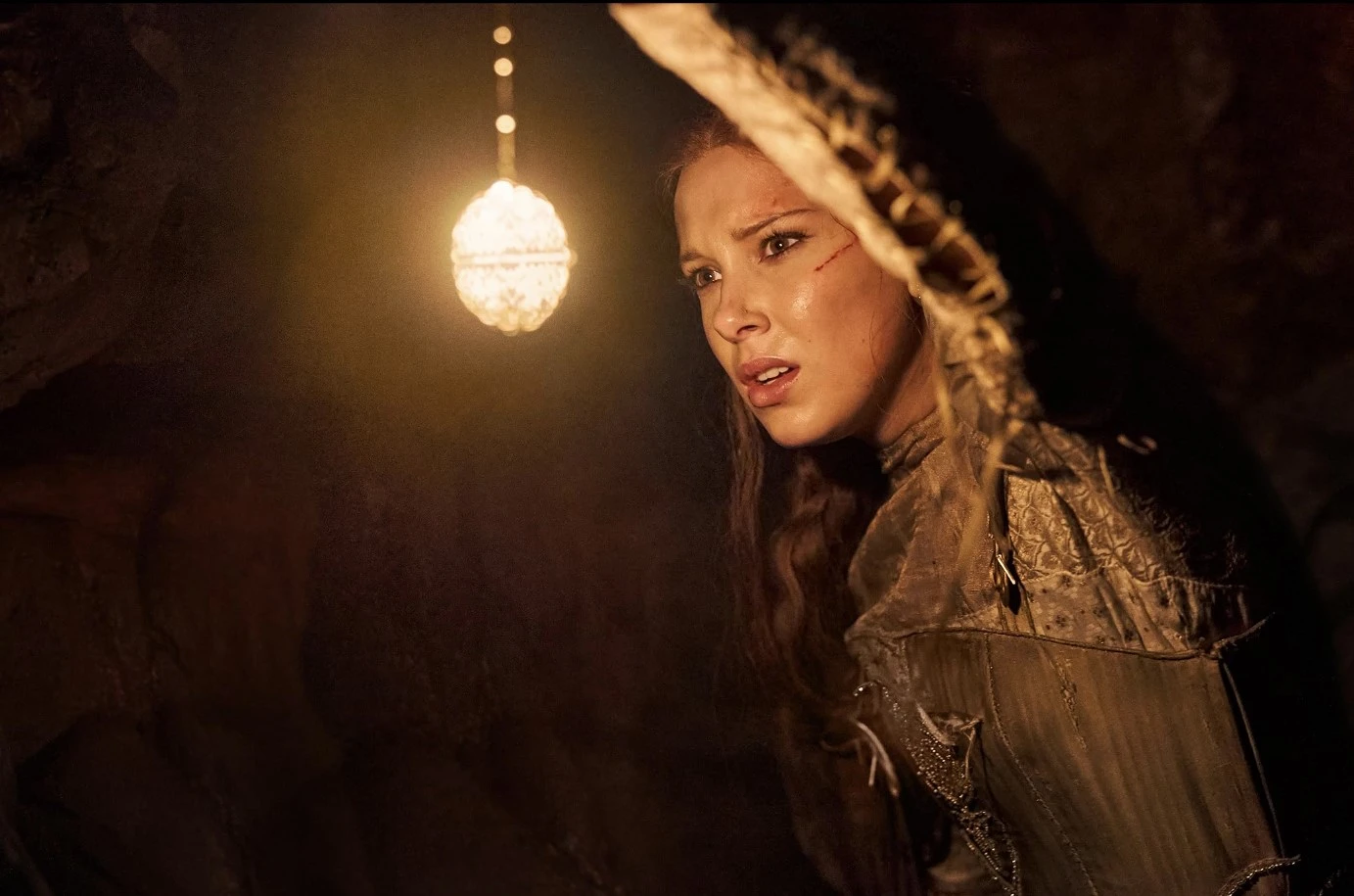 Millie Bobby Brown stands near a hanging lightbulb in a dimly-lit room.