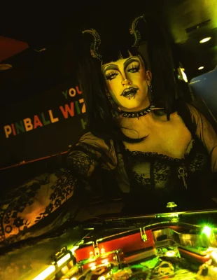 Illuminated by green and yellow lights, Nancy Raygun leans on a pinball machine.