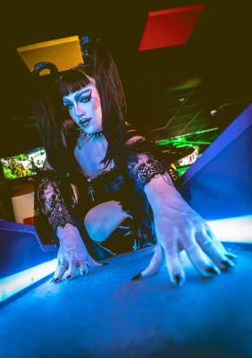 Nancy Raygun leans into a skee ball machine, illuminated by blue light.