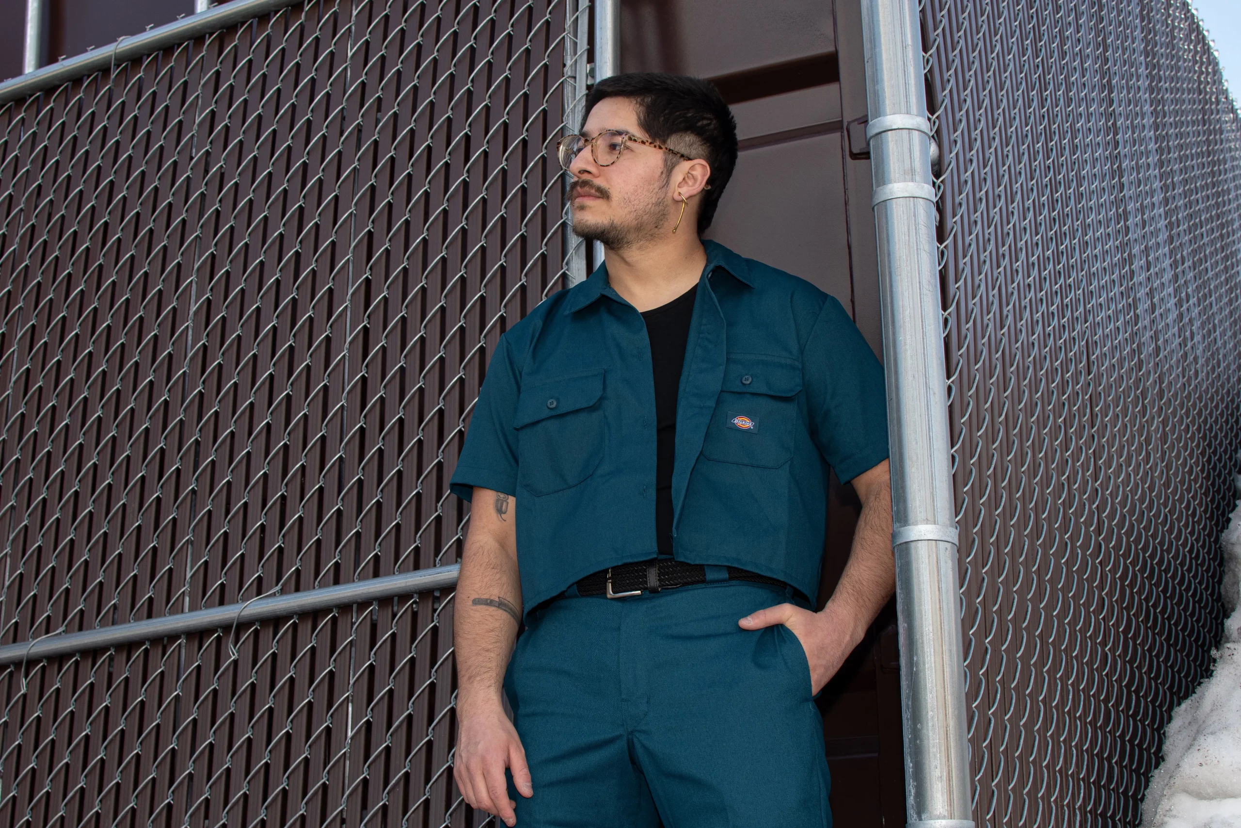 A bespectacled person with a mustache stands in front of a brown cement building encased by a chainlink fence. They wear a a blue worksuit combo from Dickies.