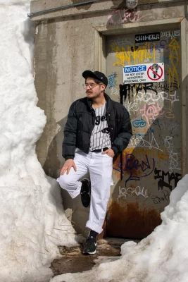 A bespectacled person with a mustache stands in front of a graffitied cement door surrounded by snow. They wear an outfit reminiscent of a Baseball player's uniform.