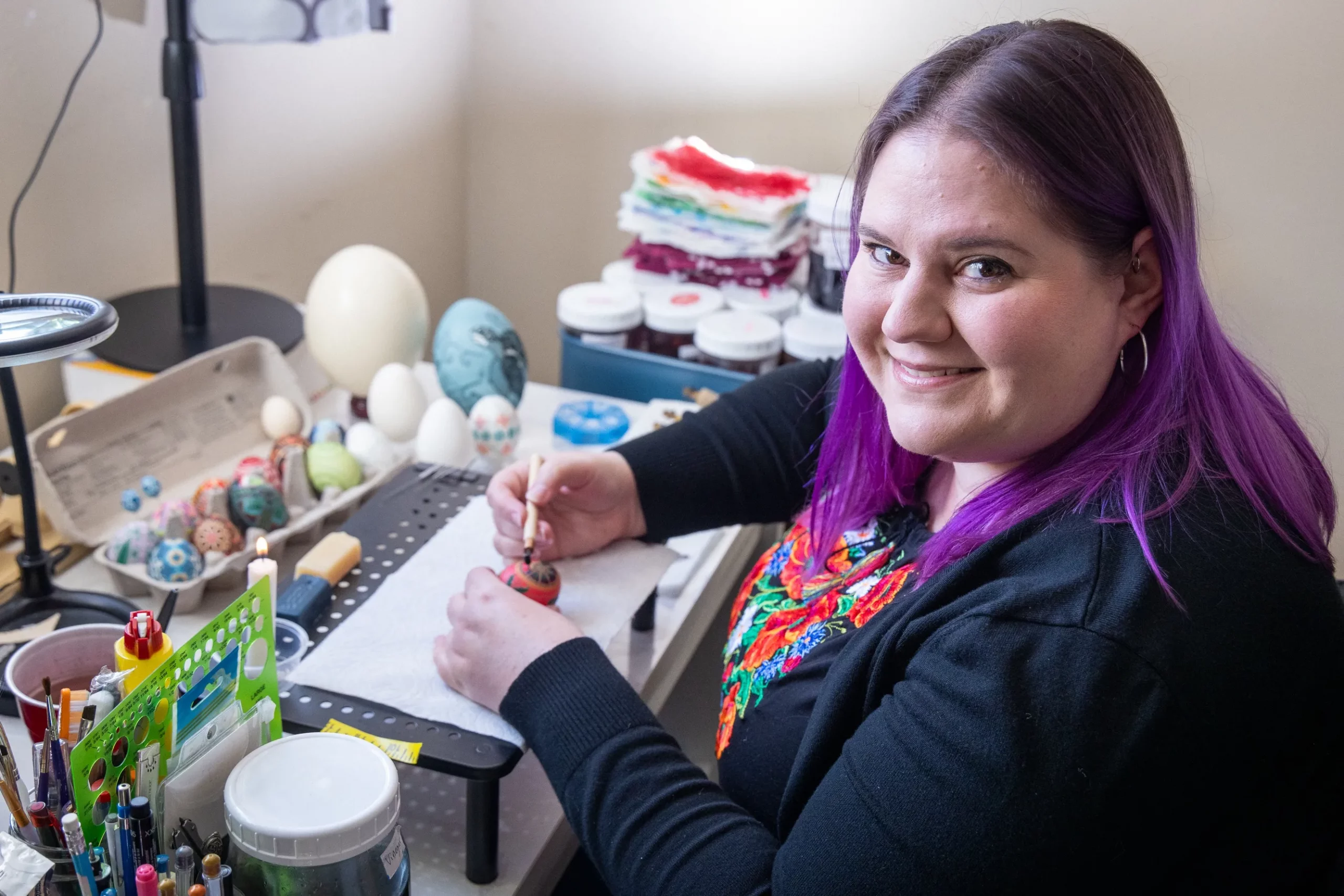A woman holds a stylus while she paints an egg.