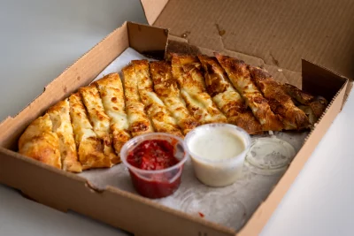 A cardboard box contains cheese pull-apart stick. Inside there is also a container of a red sauce and a white sauce.