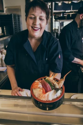 A woman in a black chef's uniform smiles as she holds a sushi bowl towards the camera.