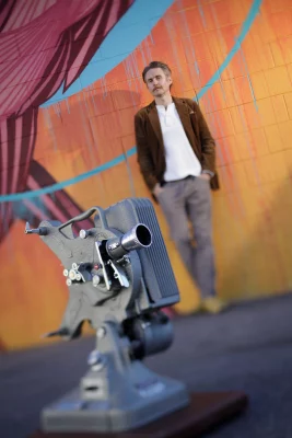 Brain Higgins leans against in background and stares at film camera in foreground.