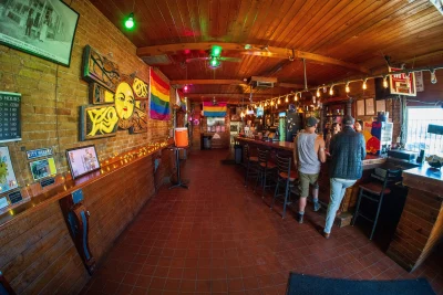 Interior of the SunTrapp. With pride flags hanging on wooden walls, patrons at the bar and warm lighting. 