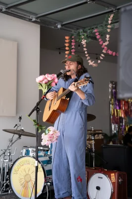 A man with long hair in a blue coverall holds an acoustic guitar while playing the harmonica in a microphone on stage.