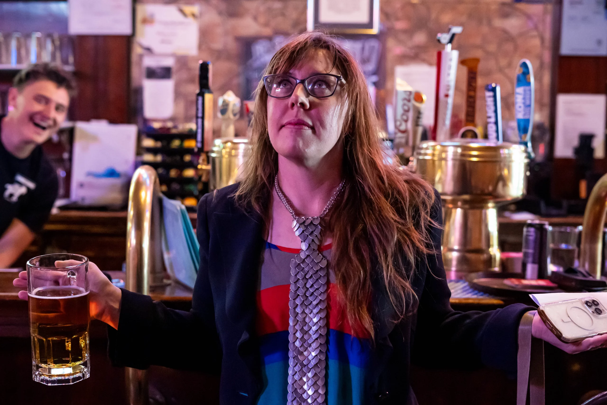 Woman holds beer and phone in each hand and looks upward.