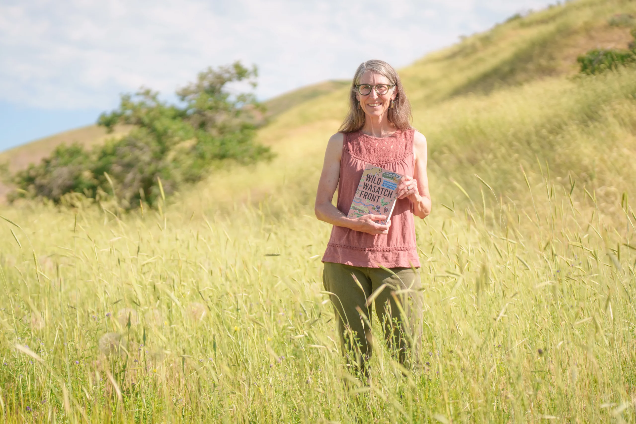 Lisa Thomspon posing with a smile holding a copy of her book "Wild Wasatch Front" in a filed of grass.