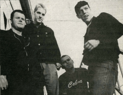 Four men in a black and white photo stands in what appears to be an outside stairwell. An open sky behind them. They wear black jackets.