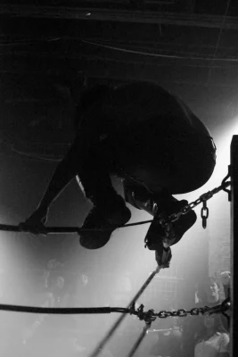 A dimly lit black and white photo of a wrestler crouched atop the ropes of the wrestling ring. 