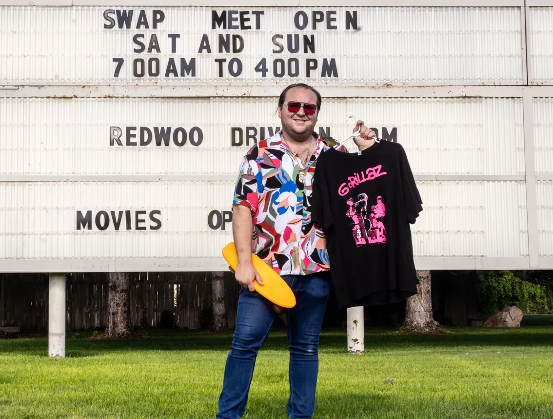 A man in sunglasses and a hawaiian shirt stands in front of a marquee smiling